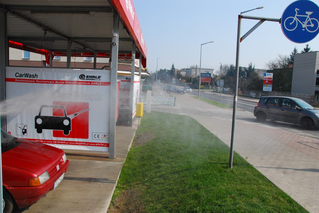 Bezdotykowa, samoobsługowa, 3-stanowiskowa myjnia samochodowa CarWash wykorzystująca technologię mycia mikroproszkiem EHRLE (Niemcy). Sfotografowanie uciążliwej mgiełki jest trudne. Wykorzystując odpowiednie oświetlenie, specjalnie podjechaliśmy samochodem, żeby zilustrować problem. Woda wydostająca się pod dużym ciśnieniem z urządzenia, trzymanego przez myjącego, odbija sią od maski pojazdu, a jej drobinki przenoszą się na pas zieleni i chodnik. Nawilżanie potwierdza trawa bujnie rosnąca między myjnią a chodnikiem   fot. Piotr P. Ruszkowski